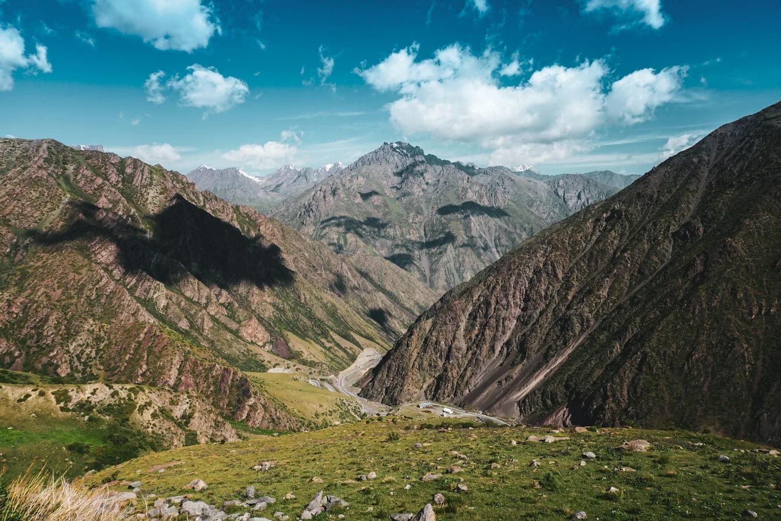 A green mountain valley in Kyrgyzstan with a road winding through the base of the peaks under a bright blue sky | BudgetBro