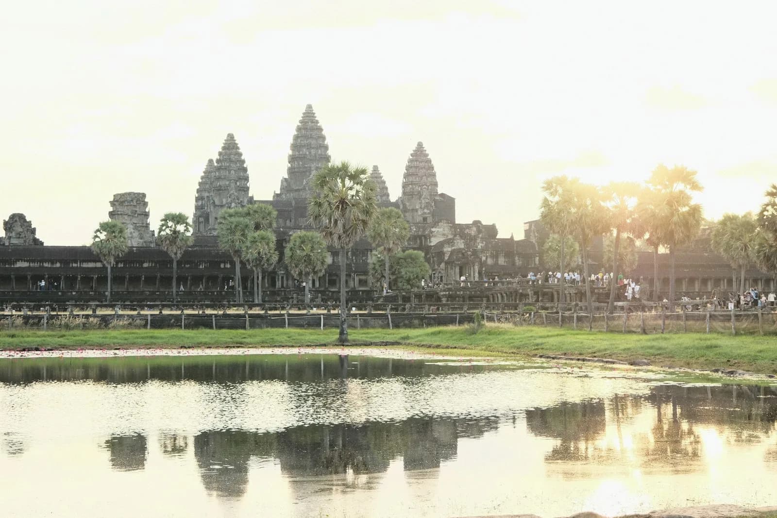 Angkor Wat temple reflected in its moat at golden hour in Siem Reap, Cambodia, with palm trees and visitors along the causeway | BudgetBro