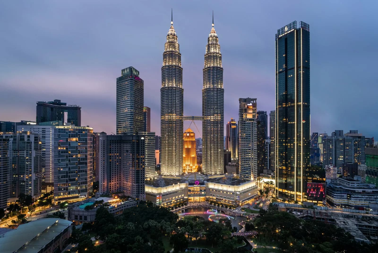 The Petronas Twin Towers lit up above the Kuala Lumpur skyline at dusk, with the KLCC park and city lights below | BudgetBro