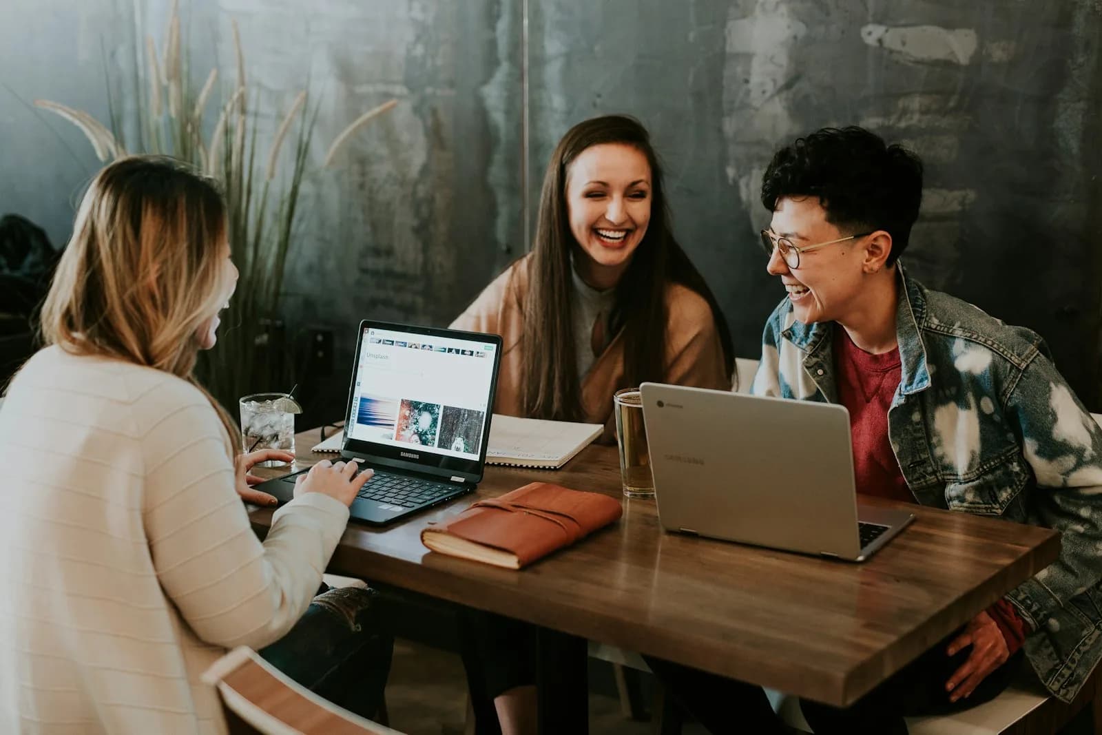 Three remote workers laughing together over their laptops at a shared table in a cafe | BudgetBro