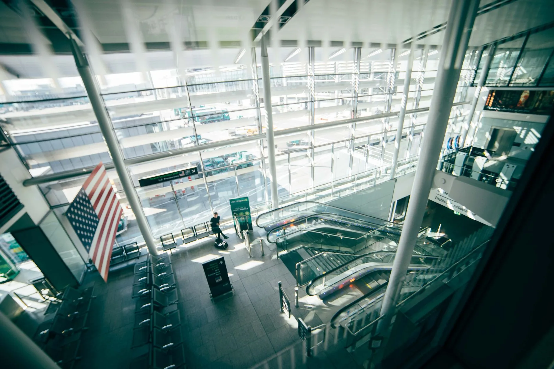 Dublin Airport travel. A bright, spacious airport terminal featuring expansive glass walls, escalators, and American flag. | BudgetBro