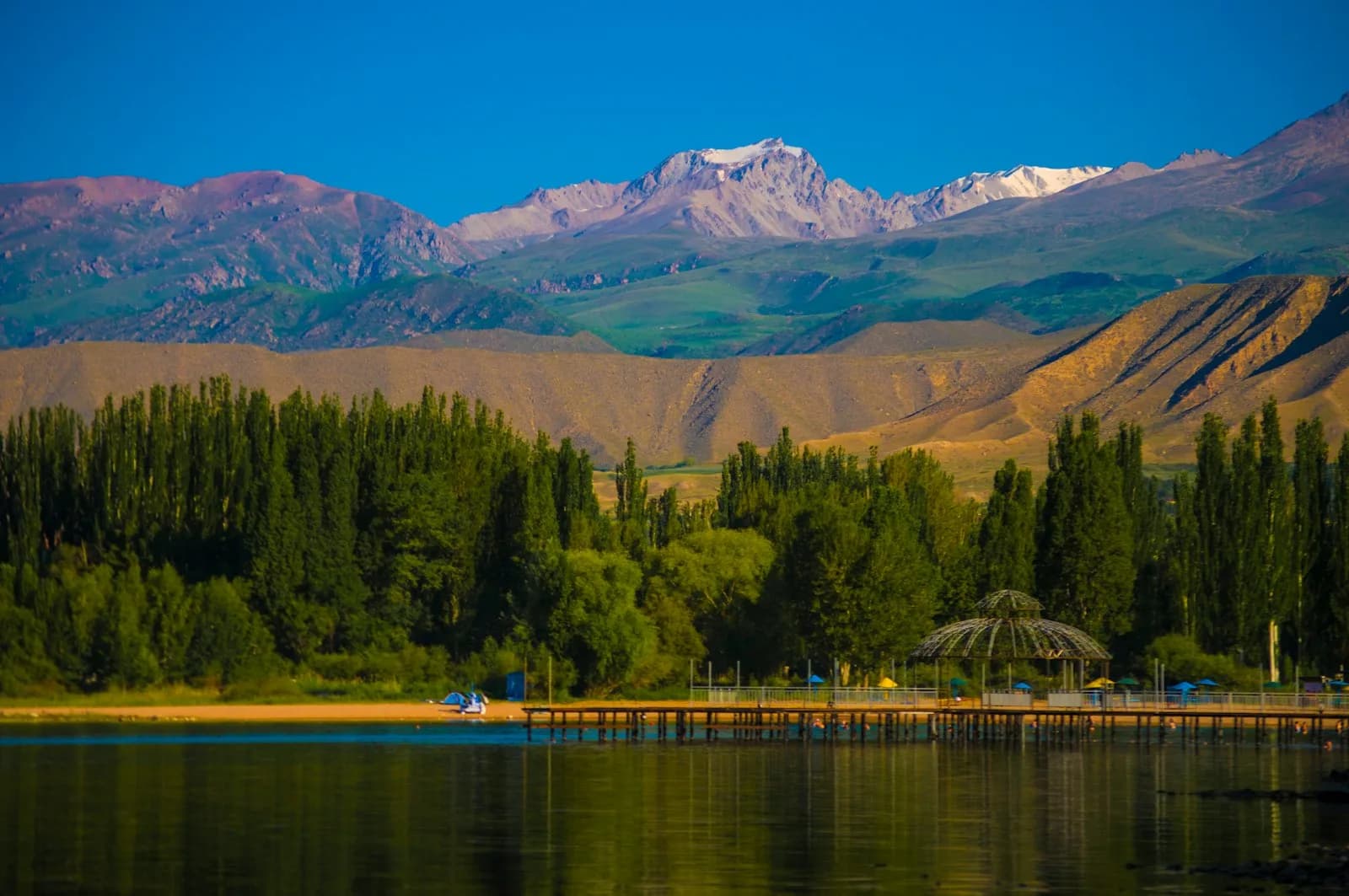 Lake Issyk-Kul in Kyrgyzstan with snow-capped mountains behind a tree-lined shore, where most World Nomad Games events are held | BudgetBro