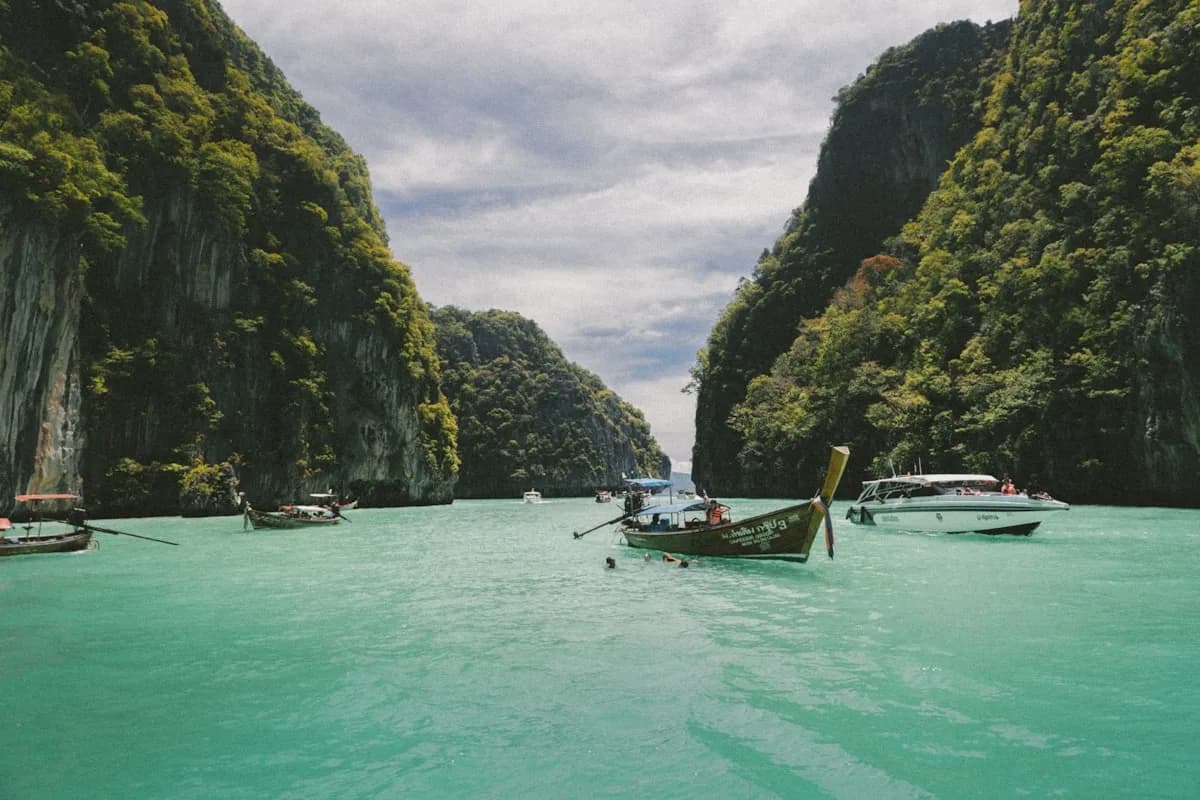 Longtail boats floating in the emerald-green lagoon at Ko Phi Phi Leh, surrounded by sheer jungle-covered limestone cliffs under an overcast sky | BudgetBro