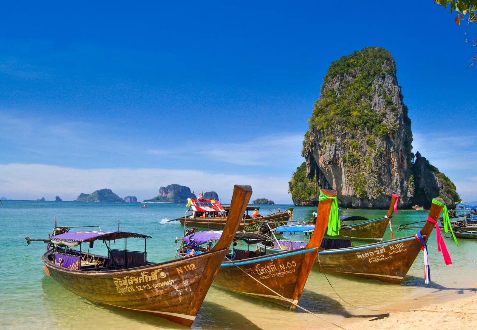 Wooden longtail boats moored on a white-sand beach at Railay in Krabi, Thailand, with limestone karst cliffs rising from turquoise water | BudgetBro